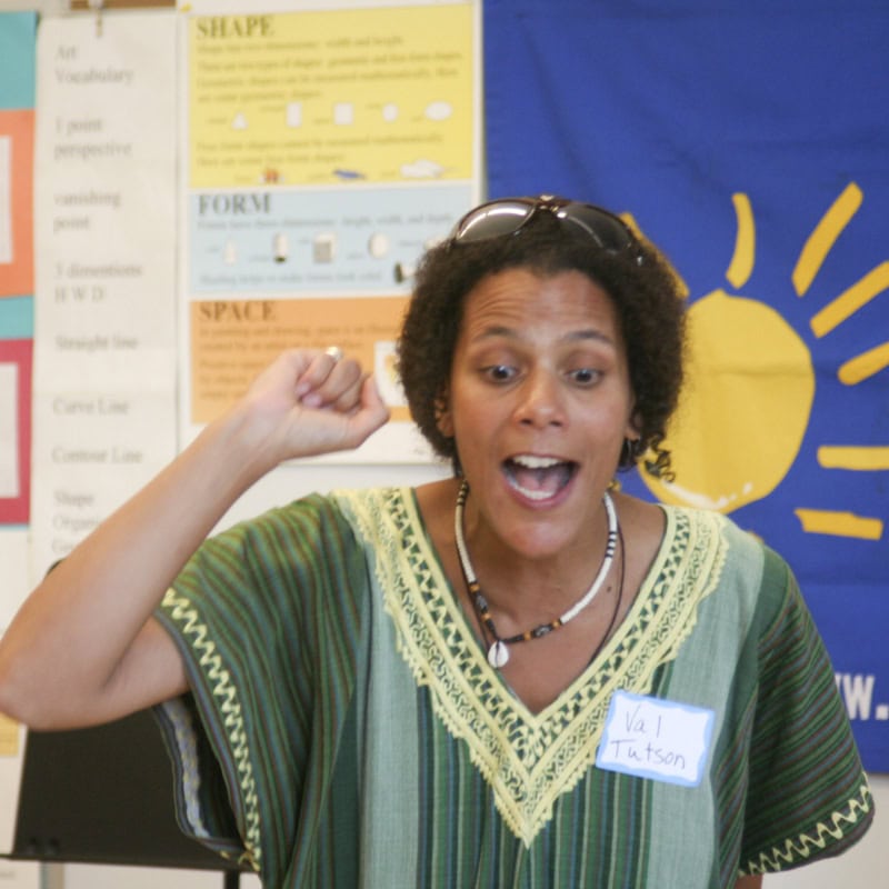 A woman wearing a green patterned shirt and a name tag stands speaking energetically, with her mouth open and one hand raised near her head. Colorful posters and a blue banner with a yellow sun are in the background.